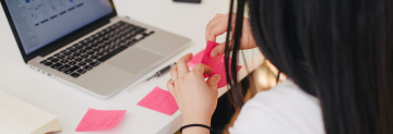 Photo of a desk over a person’s shoulder. They are peeling a post-it note from a pad. On the desk there is a laptop and several other post-it notes with writing on them.