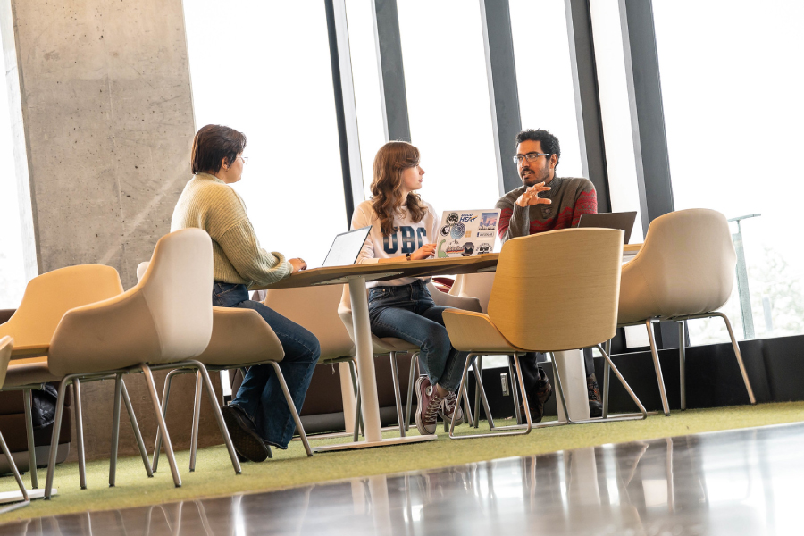 three students sitting around a table with laptops talking to each other