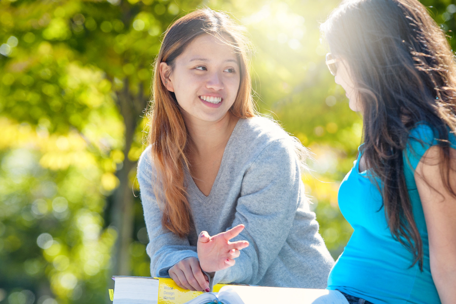 close up of two students sitting and talking to each other outside holding textbooks