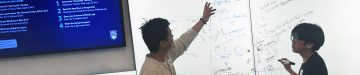 Two students writing on a white board, indoors.